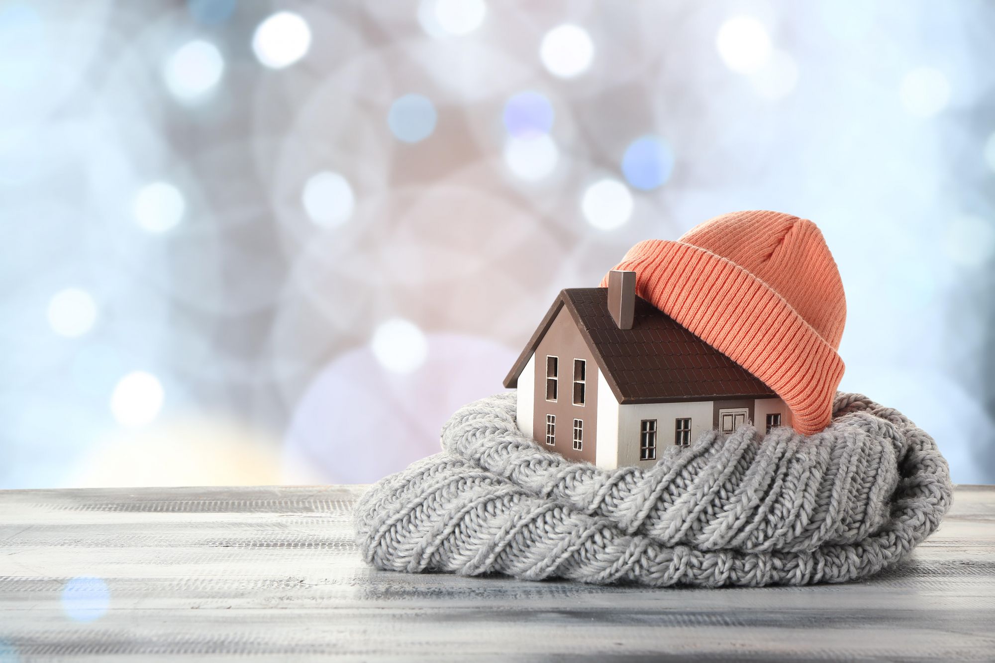 Small energy efficient house wrapped in a hat and sweater among glistening snow backdrop