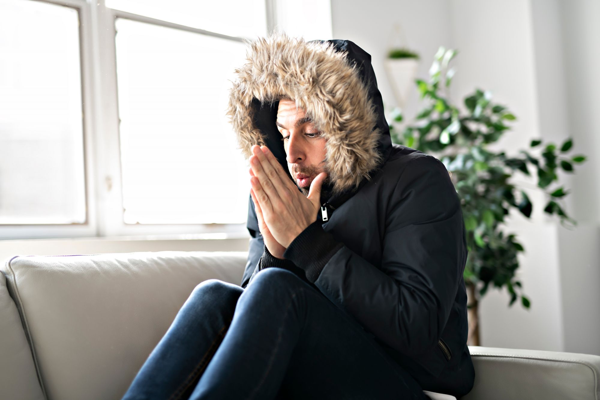 A cold man blows into his hands while wearing a coat on his couch.